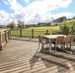 A wooden deck with a table and six chairs overlooking a green field with a house in the distance at How Beck in Hawkshead