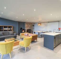 An open living room with yellow chairs a gray sofa a brown sofa a glass coffee table a wooden dining table a brick fireplace and a kitchen island at Gillside in Millbeck