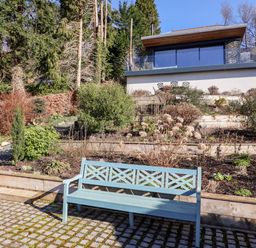 A blue bench on a paved area in front of terraced garden beds with bushes and a modern house in the background at Gillside in Millbeck