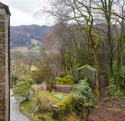 A stone pathway with bushes and mossy trees next to a stone building and a small stone structure in a wooded area at Silver Howe in Grasmere