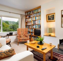 A living room with patterned armchairs a wooden coffee table a bookshelf and a window showing trees and hills at The Hayloft in Ambleside