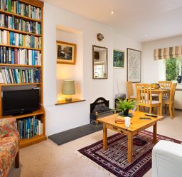 A living room with a wooden bookshelf filled with books a small TV a wooden coffee table with plants and books a dining table with chairs and windows at The Hayloft in Ambleside