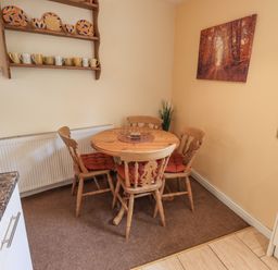 A kitchen dining area with a round wooden table and four chairs with cushions a wall shelf with plates and mugs and a framed picture on the wall at Hideaway Cottage in Ambleside