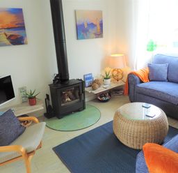 A living room with a wood stove, blue sofas, a wicker coffee table, a wooden chair with a pillow, and paintings on the walls at Bryn Mor in Church Bay