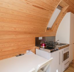 A small kitchen with a white table and chair a stove with a kettle a dish rack a sink and a wooden wall at Halmore Pod Halmore near Berkeley