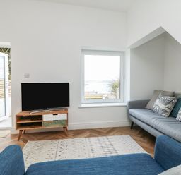 A living room with a blue chair a gray sofa a rug a TV on a wooden stand and a window with a view at Through The Porthole in Falmouth