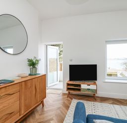 A living room with a wooden sideboard a round wall mirror a television on a stand and a door leading outside at Through The Porthole in Falmouth