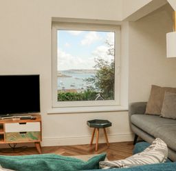 A living room with a television on a wooden stand a small table a gray sofa with cushions and a view of boats on water through a window at Through The Porthole in Falmouth