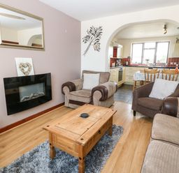 A living room with armchairs a coffee table and a wall-mounted fireplace next to a kitchen at Rhiangwyn Cottage in Llanrhyddlad near Cemaes Bay