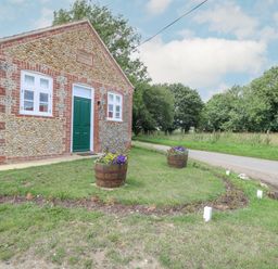 A small stone building with a green door and two white windows next to a road with flower pots on grass at Wensum Spring in Horningtoft near Fakenham