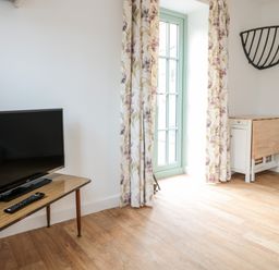 A room with a television on a wooden table next to floral curtains and a folding table against the wall at Ostlers in Mitcheldean