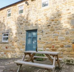 A stone exterior wall with a blue door and four windows with a wooden picnic table holding a wine bottle and glasses in front at Todridge Barn in Corbridge