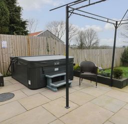 An outdoor patio area with a hot tub brown wicker chair and green grass at Big Azzles in Tyn-y-Gongl near Benllech