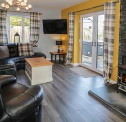A living room with black leather sofas a wooden coffee table a wall-mounted television and a wood-burning stove at Big Azzles in Tyn-y-Gongl near Benllech