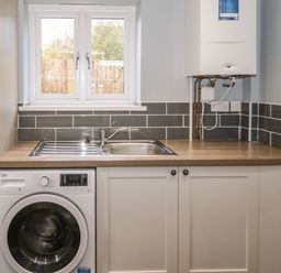 A utility room with a washing machine under a wooden countertop with a sink and a boiler on the wall at Big Azzles in Tyn-y-Gongl near Benllech