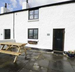 Exterior view of a white stone cottage with black doors and windows a wooden picnic table on the stone patio at Menai Cottage in Brynsiencyn