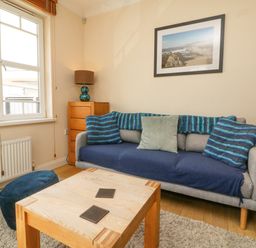 A living room with a grey sofa with blue and green cushions a wooden coffee table a chest of drawers with a lamp and a framed picture on the wall at Beach Lodge in Woolacombe