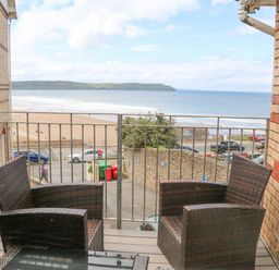 A balcony with two wicker chairs and a glass table overlooking a beach and ocean at Beach Lodge in Woolacombe