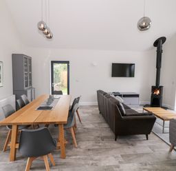 A living room and dining area with a wooden dining table and gray chairs a brown sofa a TV mounted on the wall and a wood stove at Pen Y Cefn in Pen Y Cefn