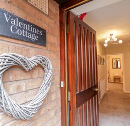 An open wooden front door with a heart-shaped wreath and a sign reading Valentines Cottage leading to a hallway with carpet and stairs at Valentines Cottage Stratford-Upon-Avon