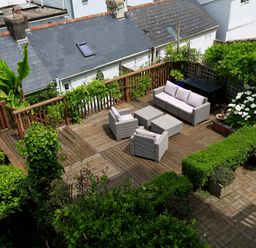 A wooden outdoor patio with cushioned seating and surrounded by various green plants and bushes at Armorel House in Dartmouth
