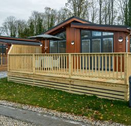 A wooden cabin with a fenced deck and patio furniture surrounded by trees at Otter Lodge in Gargrave