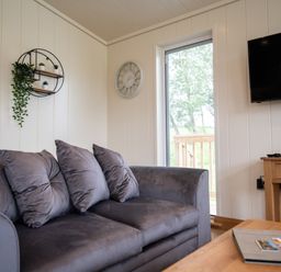 A living room with a gray sofa pillows a wall clock a wall shelf with plants a wooden table and a wall-mounted TV at Otter Lodge in Gargrave