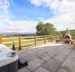 An outdoor patio with a hot tub and a bottle of wine on a tray and a barbecue oven with picnic tables and countryside views at The Stables At Cae Gwyn in Ruthin