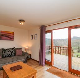 A living room with a sofa pillows and a coffee table near sliding glass doors leading to a wooden deck and countryside at The Willow Lodge near Lyme Regis