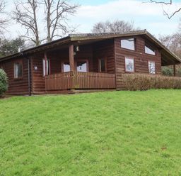 A wooden cabin with a porch and multiple windows on a grassy slope near trees at The Oak Lodge near Lyme Regis