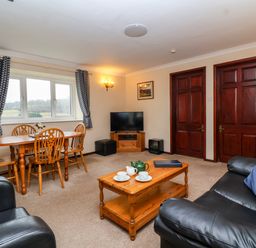 A living room with a wooden dining table and chairs a television on a wooden stand and black leather sofas at The Hayloft Cottage Apartment near Lyme Regis
