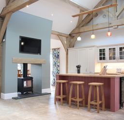 A kitchen with an island and three stools under pendant lights and a stove below a mounted television at The Anchorage @ Nables Farm in Upper Seagry