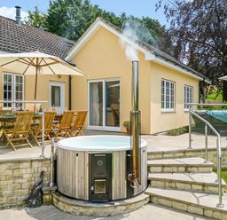 An outdoor patio area with wooden dining chairs and table under umbrellas and a round hot tub at The Anchorage @ Nables Farm in Upper Seagry