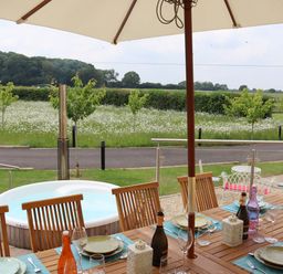 An outdoor dining area with a wooden table set with plates and glasses under an umbrella next to a round hot tub overlooking a field at The Anchorage @ Nables Farm in Upper Seagry