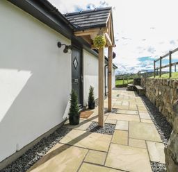 A paved pathway alongside a white building with a black door and potted plants near a wooden canopy and stone wall at Ty Cuddfan in Llangoed