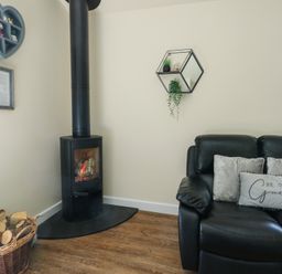 A corner with a black wood burning stove next to a basket of firewood and a black sofa with cushions at Ty Cuddfan in Llangoed