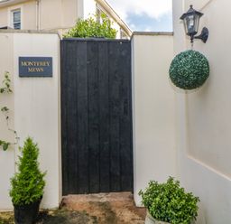 A black wooden gate in a white wall with a hanging lamp and plants at Monterey Mews in Torquay