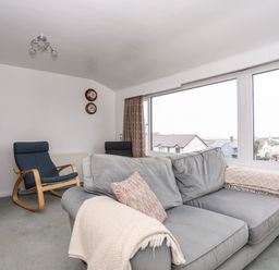 A living room with a gray sofa blue rocking chairs and large windows overlooking houses and the sea at The Little Dolphins in Rhosneigr