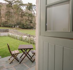 A wooden table and two chairs on a stone patio overlooking a green lawn with a stone wall and trees at Oak Tree Cottage in Coniston