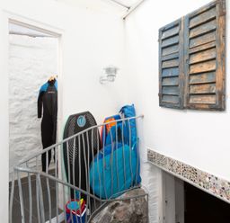 A small indoor space with a wetsuit hanging in a doorway a bodyboard and blue bags leaning against the wall a bucket with beach toys on the floor and rustic wooden shutters on the wall at The Sail Loft in St Ives