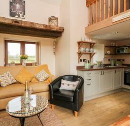 A small living area with a sofa, armchair, glass coffee table and kitchen with wooden shelves at Tegfan Barn Pant-y-dwr near Rhayader
