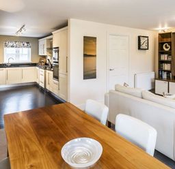 An open plan living space with a wooden dining table and white chairs a white sofa a built in shelving unit a wall clock and a kitchen with cream cabinets at The Golf Apartment in Kendal