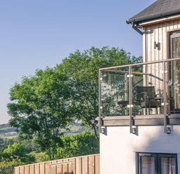 A balcony with two chairs and a small table on a wooden house overlooking trees and distant hills at Swallows Dip in Honiton
