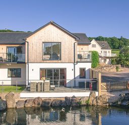 A two story building with wooden and white exterior walls overlooking a pond with rocks and outdoor furniture on a patio at Swallows Dip in Honiton