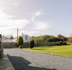 A gravel driveway next to a lawn with trees and houses behind a wooden fence at Penllyn in Newborough