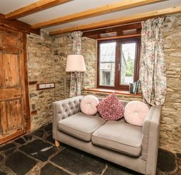 A room with stone walls wooden door window with floral curtains a gray sofa with cushions and a floor lamp at Pant Y Rhedyn Abbeycwmhir near Rhayader
