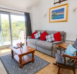A living room with a gray sofa with red and checkered cushions a wooden rocking chair a wooden coffee table on a patterned rug and sliding glass doors at Higher Bumsley Barn in Parracombe