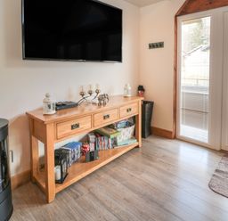 A wooden console table with decorative items and DVDs under a wall mounted TV next to glass doors with blinds at Treetops in Otterburn