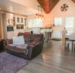 An open-plan living room with brown leather sofas and a purple rug next to a kitchen with white cabinets and a dining area with a wooden table and chairs at Treetops in Otterburn