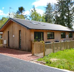 A wooden single-story house with a fenced patio and trees in the background at Broken-Sky Lodge in Otterburn